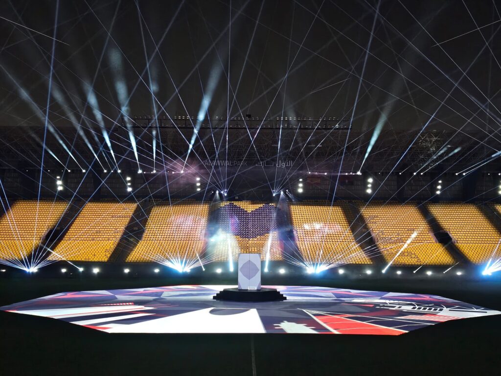 Stadium stage with geometric floor visuals and white laser beams radiating across the venue