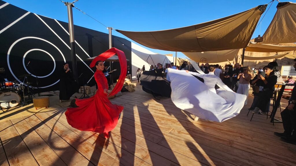 Dancers in flowing red and white costumes performing at an outdoor desert event with live music and audience